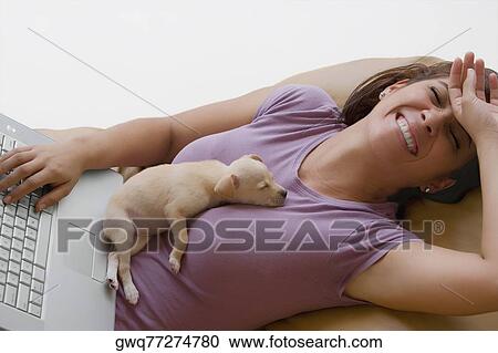 Young woman using a laptop and laughing with a puppy lying on her View Large Photo Image Stock Image - Young woman using a laptop and laughing with a puppy lying on her. Fotosearch
