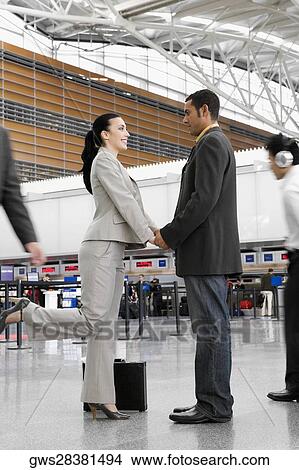Picture - Businessman and a businesswoman standing at an airport with holding each other's hands. Fotosearch