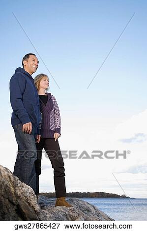 Stock Photo - Low angle view of a mature couple standing on a rock and smiling. Fotosearch
