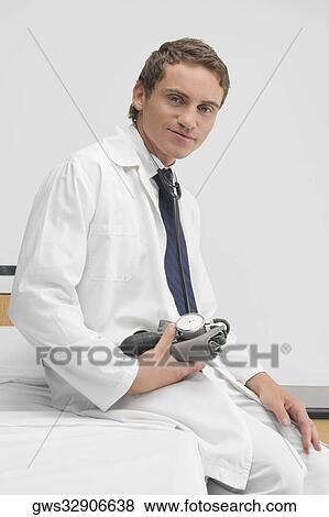 Portrait of a male doctor sitting on the bed and holding a blood pressure gauge View Large Photo Image Stock Photo - Portrait of a male doctor sitting on the bed and holding a blood pressure gauge. Fotosearch
