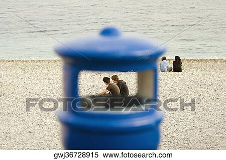 Couple sitting on the beach viewed through a garbage bin, Baie Des Anges, Nice, Provence-Alpes-Cote D'Azur, France View Large Photo Image Stock Photography - Couple sitting on the beach viewed through a garbage bin, Baie Des Anges, Nice, Provence-Alpes-Cote D'Azur, France. Fotosearch
