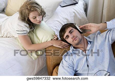Stock Image - High angle view of a young man listening to an MP3 player with a young woman lying on the bed. Fotosearch