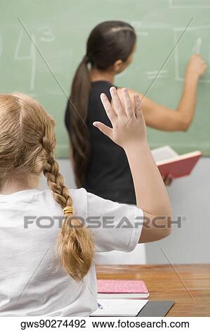 Stock Image - Rear view of a schoolgirl with her hand raised in a classroom. Fotosearch