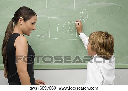 Schoolboy writing on a blackboard and his teacher looking at him View Large Photo Image Stock Photo - Schoolboy writing on a blackboard and his teacher looking at him. Fotosearch