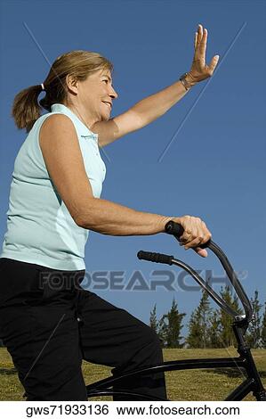 Stock Photograph - Side Profile of a senior woman cycling and waving her hand. Fotosearch