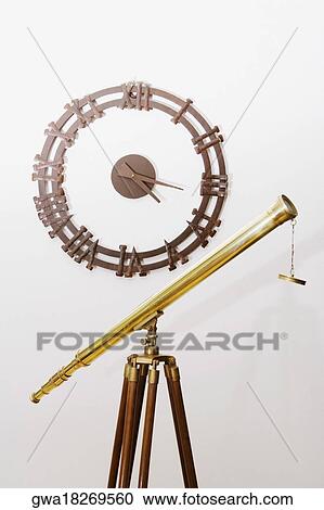 Stock Image - Telescope on a tripod with a clock on a wall. Fotosearch