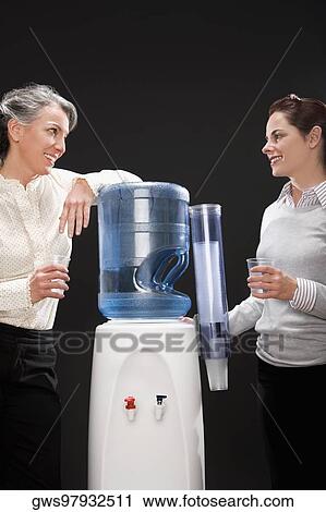 Stock Image - Two businesswomen gossiping near a water cooler. Fotosearch