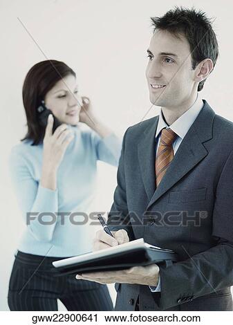 Stock Image - Businessman holding a personal organizer with a businesswoman talking on a mobile phone behind him. Fotosearch