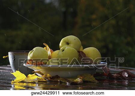 Stock Image - Close-up of a bowl of apples with cups and fallen leaves on the table. Fotosearch