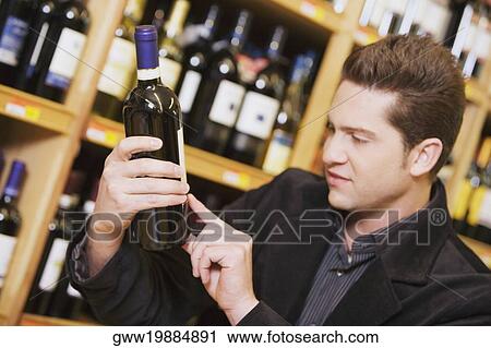 Stock Image - Close-up of a young man reading a label of a wine bottle in a liquor store. Fotosearch