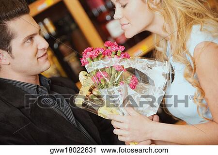 Stock Image - Close-up of a young woman smelling a bouquet of flowers and a young man looking at her. Fotosearch