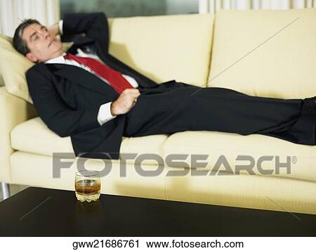 Stock Image - Glass of whiskey on the table with a businessman lying on the couch. Fotosearch