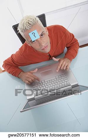 Stock Image - High angle view of a young man in front of a laptop with an adhesive note on his forehead. Fotosearch