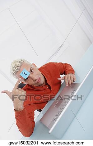 Stock Image - High angle view of a young man using a laptop with an adhesive note on his forehead. Fotosearch