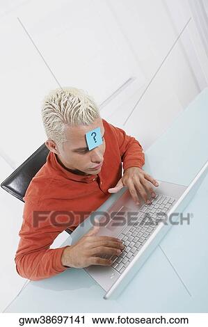 Stock Image - High angle view of a young man using a laptop with an adhesive note on his forehead. Fotosearch