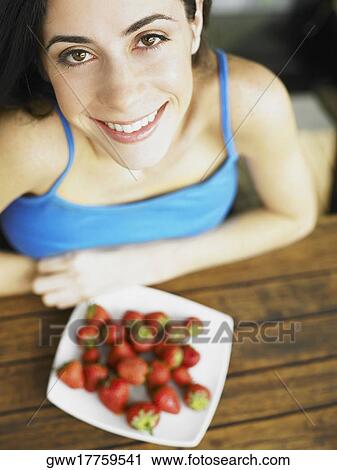 Stock Image - High angle view of a young woman smiling with a plate of strawberry in front of her. Fotosearch