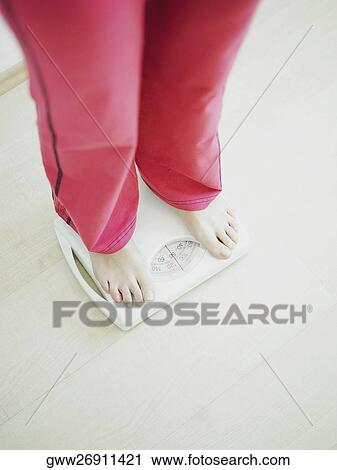 Stock Image - Low section view of a woman standing on a weighing scale. Fotosearch
