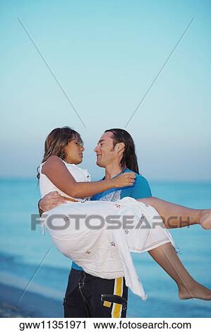 Stock Image - Mid adult man carrying a young woman on the beach. Fotosearch