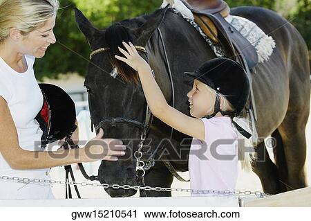 Stock Image - Mid adult woman and her daughter touching a horse. Fotosearch