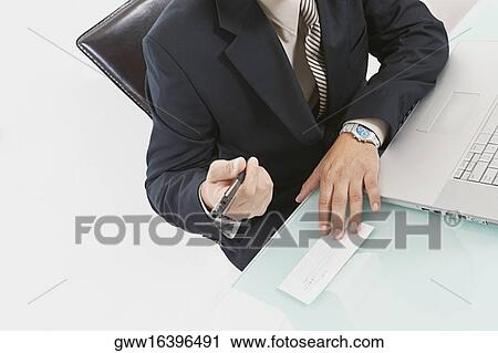 Stock Image - Mid section view of a businessman sitting in front of a laptop and holding a pen and a check. Fotosearch