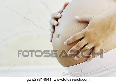 Stock Image - Mid section view of a pregnant woman touching her abdomen. Fotosearch