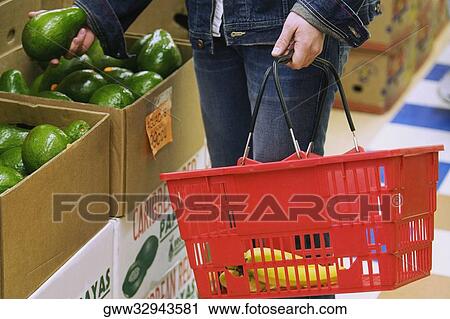 Stock Image - Mid section view of a woman holding a mango and shopping basket. Fotosearch