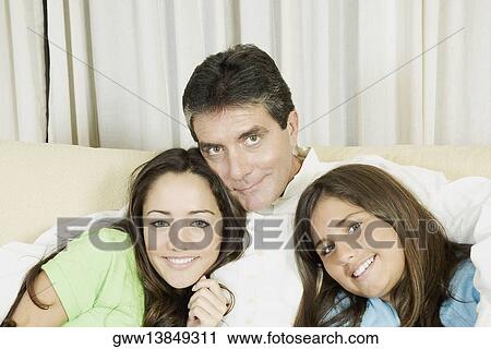 Stock Image - Portrait of a mature man sitting with his two daughters on a couch and smiling. Fotosearch