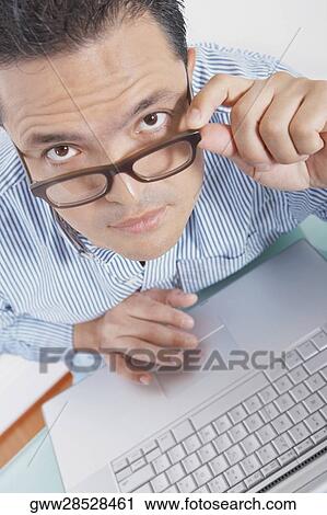 Stock Image - Portrait of a mid adult man using a laptop and adjusting his eyeglasses. Fotosearch