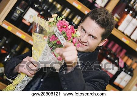 Stock Image - Portrait of a young man holding a bouquet of flowers and pointing forward in a liquor store. Fotosearch