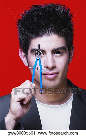 Stock Image - Portrait of a young man holding a drawing compass in front of his eye. Fotosearch