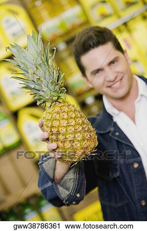 Stock Image - Portrait of a young man holding a pineapple in a supermarket. Fotosearch