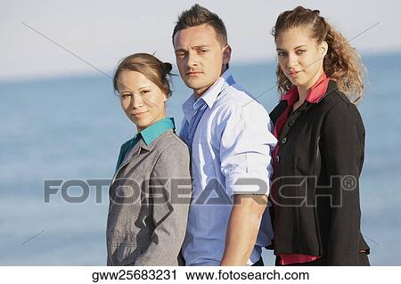 Stock Image - Portrait of a young man with two young women standing on the beach. Fotosearch