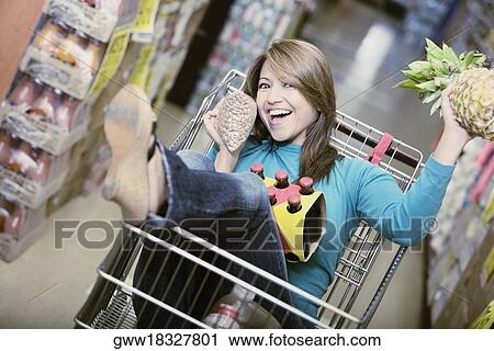 Stock Image - Portrait of a young woman sitting in shopping cart and holding a pineapple. Fotosearch
