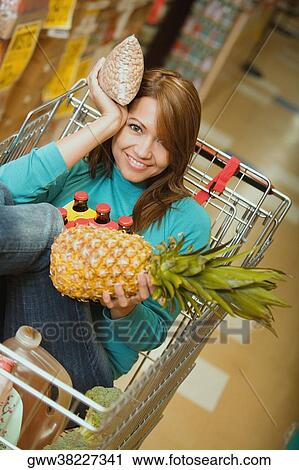 Stock Image - Portrait of a young woman sitting in shopping cart and holding a pineapple and a packet. Fotosearch