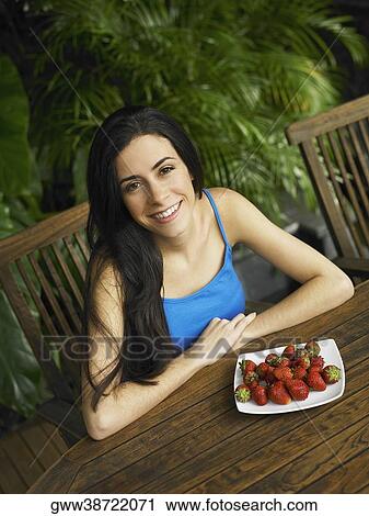 Stock Image - Portrait of a young woman smiling with a plate of strawberries in front of her. Fotosearch