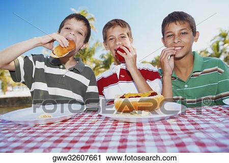 Stock Image - Portrait of two boys and a teenage boy sitting at a table and eating hot dogs and an apple. Fotosearch