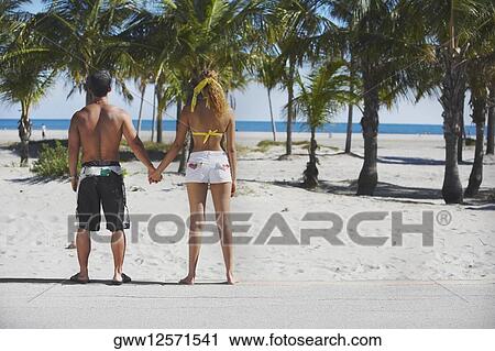 Rear view of a couple standing and holding hands on the beach View Large Photo Image Stock Image - Rear view of a couple standing and holding hands on the beach. Fotosearch