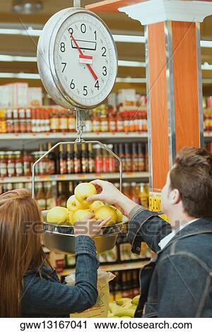 Stock Image - Rear view of a young couple weighing apples on a weighing scale in a supermarket. Fotosearch