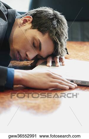 Stock Image - Side profile of a businessman resting his head on a desk in front of a laptop. Fotosearch