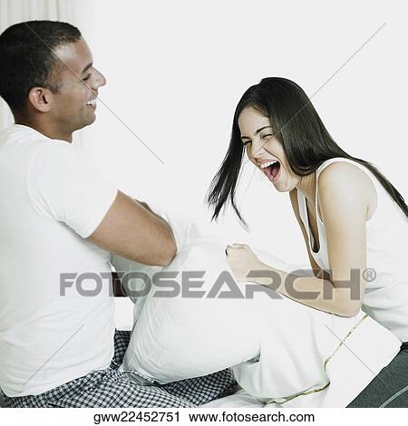 Stock Image - Side profile of a mid adult man with a young woman having a pillow fight on the bed. Fotosearch