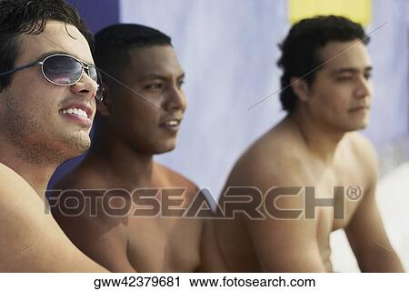 Stock Image - Side profile of a young man wearing sunglasses and sitting with his friends. Fotosearch