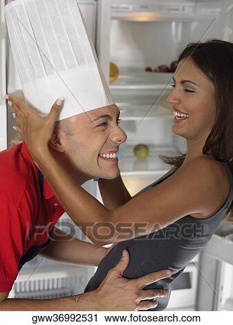 Stock Image - Side profile of a young woman holding a chef hat on a young man's head. Fotosearch
