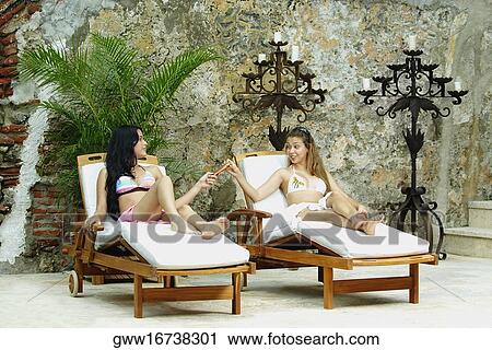 Stock Image - Two young women sitting on a lounge chair and talking to each other. Fotosearch