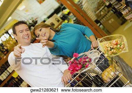 Stock Image - Young woman and a mid adult man standing in a supermarket and pointing forwards. Fotosearch