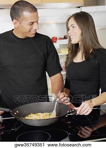 Stock Image - Young woman looking at a mid adult man cooking food and smiling. Fotosearch