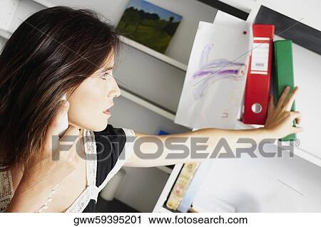 Close-up of a businesswoman picking up a ring binder from a shelf and using a telephone View Large Photo Image Stock Image - Close-up of a businesswoman picking up a ring binder from a shelf and using a telephone. Fotosearch