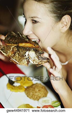 Stock Image - Close-up of a young woman eating a grilled fish. Fotosearch