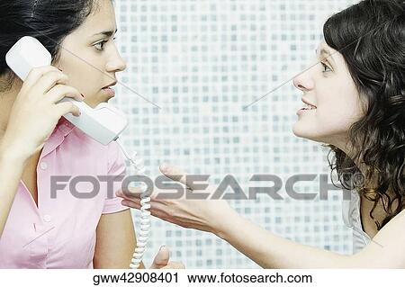Stock Image - Close-up of a young woman using a telephone with her friend sitting in front of her. Fotosearch
