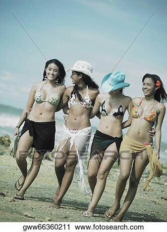 Stock Image - Four young women standing with their arms around each other walking on the beach. Fotosearch