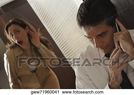 Mature man secretively talking on a mobile phone with his wife trying to listen in View Large Photo Image Stock Image - Mature man secretively talking on a mobile phone with his wife trying to listen in. Fotosearch